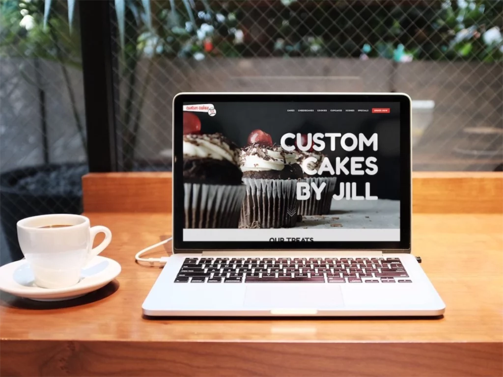 A open laptop on a cafe table showing the Custom Cakes by Jill website